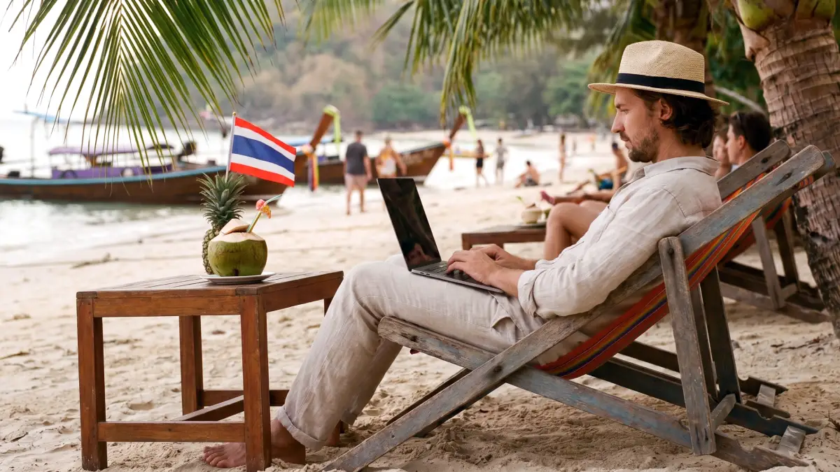 Un digital nomade assis dans un transat en bois sur une plage en Thaïlande, travaillant sur son ordinateur portable à l'ombre des palmiers. À côté de lui, une table basse avec une noix de coco à boire surmontée d'un petit drapeau thaïlandais, avec des bateaux traditionnels long-tail en arrière-plan.